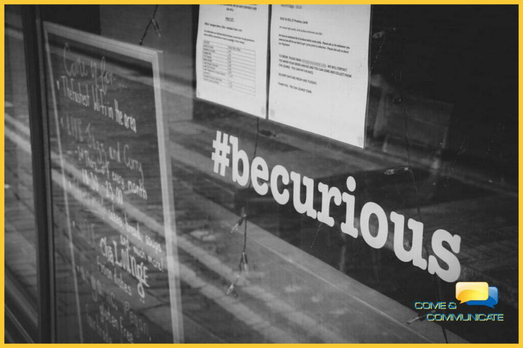 Frase #becurious puesta en vinilo en la ventana de un local junto con una pizarra negra.