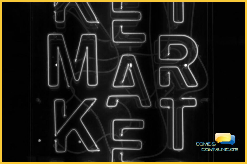 Imagen artística en blanco y negro con un borde amarillo que muestra un letrero de neón brillante con la palabra 'MARKET' dispuesta verticalmente. El diseño tiene un estilo retro-moderno con cables visibles sobre un fondo oscuro, y el logotipo de Come & Communicate aparece en la esquina inferior derecha.