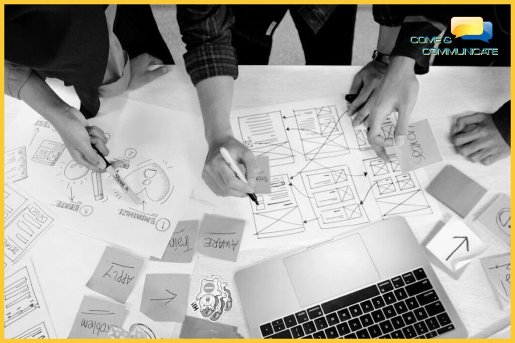 Vista cenital de un equipo colaborando en el diseño de una interfaz de usuario (UX/UI). Manos dibujan en bocetos de wireframes de papel y utilizan notas adhesivas dispersas con texto legible que incluye 